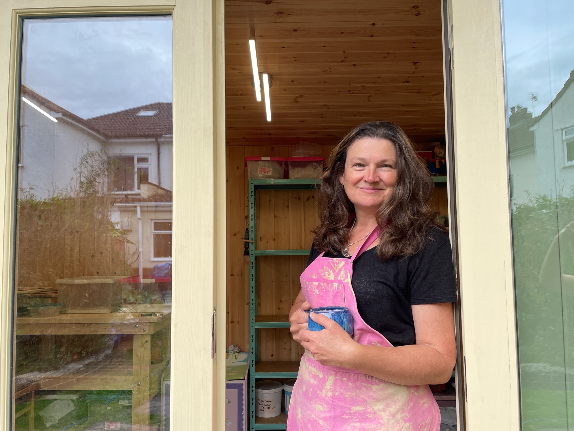 Ellie smiling in the doorway of her new garden studio, wearing a pink glaze-splashed apron and holding a mug