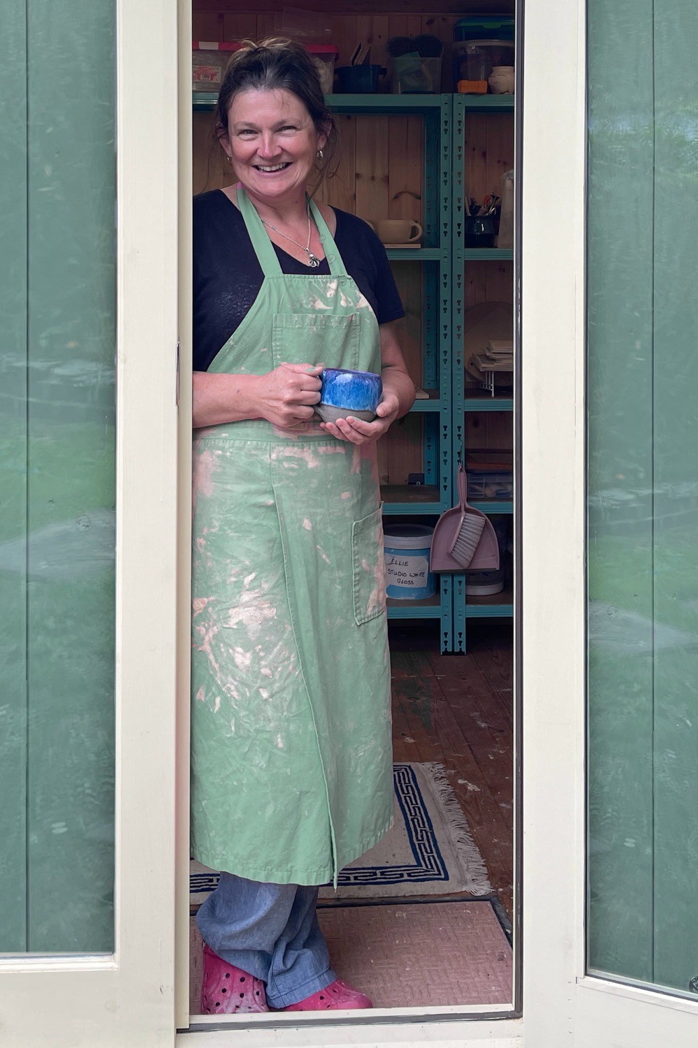 Ellie smiling in the doorway of her sage-green garden studio, wearing a clay-dusted apron and holding a blue mug