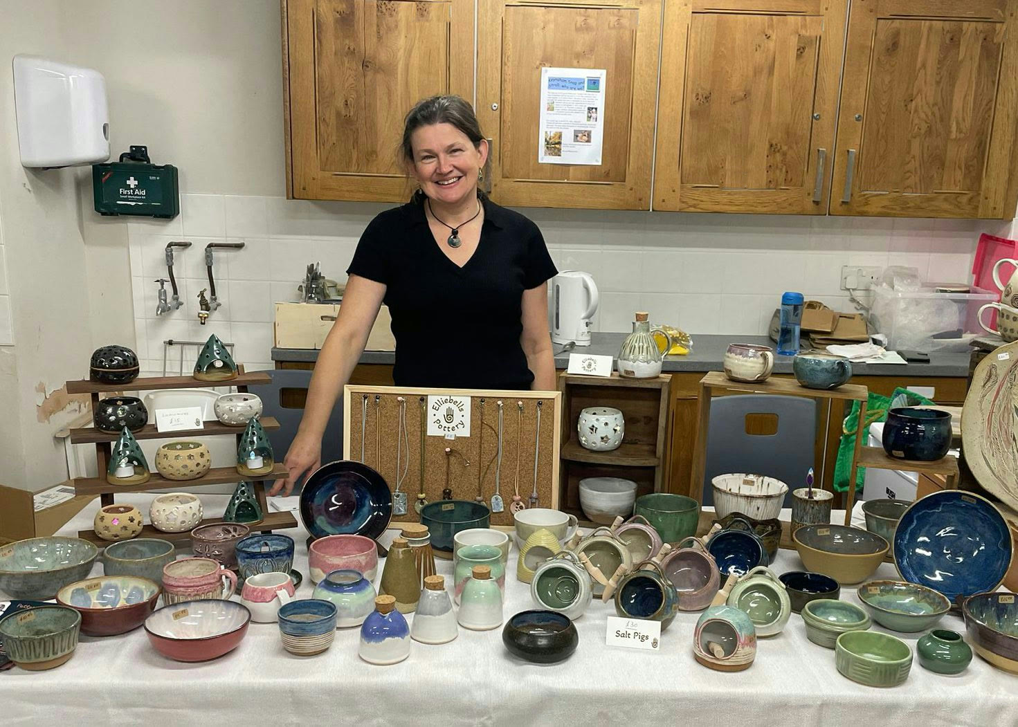 Ellie smiling behind her Elliebells Pottery stall, laid out with bowls, mugs, salt pigs, tealight lanterns and pendants