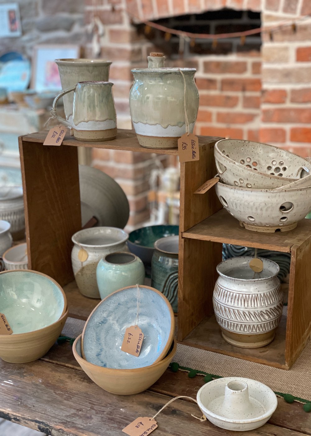 Sage drip-glaze jug and lidded jar on a wooden crate, above carved vases and pale blue bowls with brown price tags