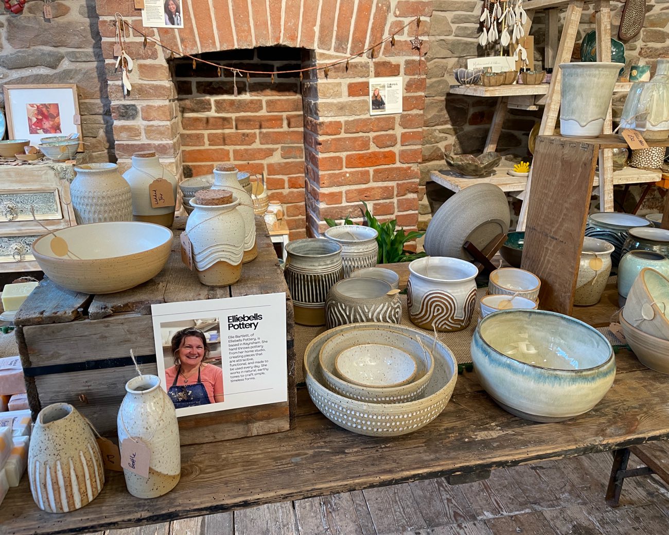 An Elliebells Pottery display on a rustic table: carved and speckled vases, nesting bowls and Ellie's maker card