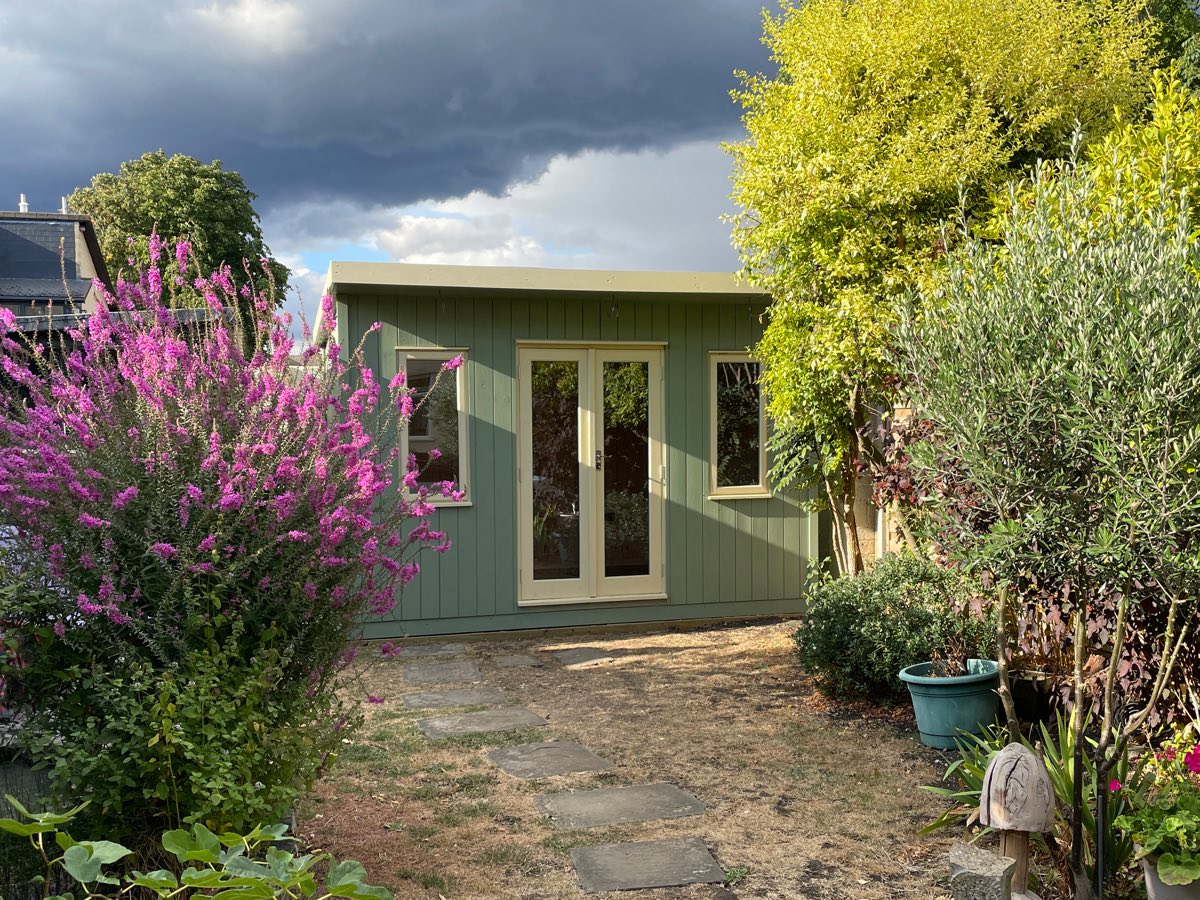 The finished sage-green garden studio with cream double doors, framed by flowering shrubs