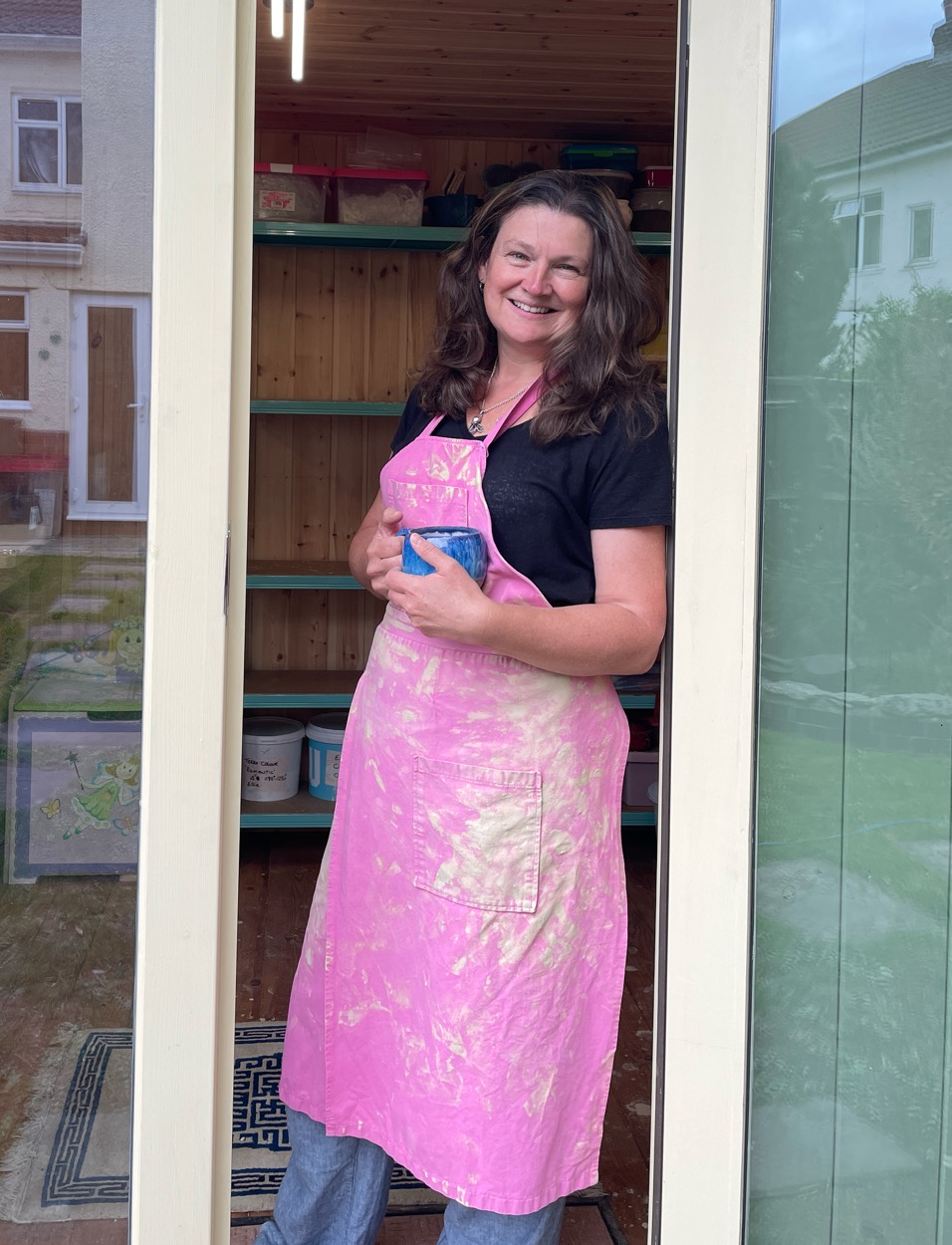 Ellie in a pink glaze-splashed apron, smiling in the open doorway of her new studio with a mug in hand