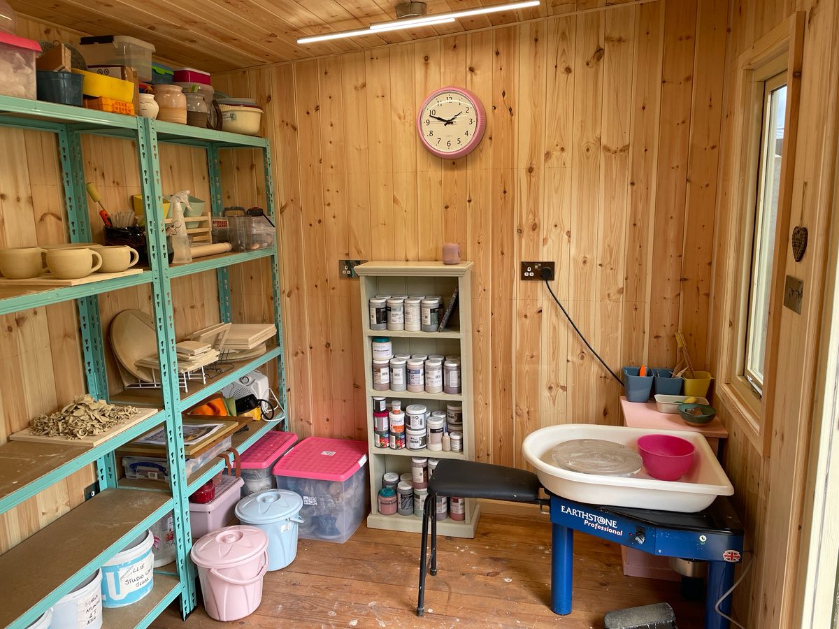 Inside the pine-lined studio: green shelving full of pots, a clock, and the potter's wheel by the wall