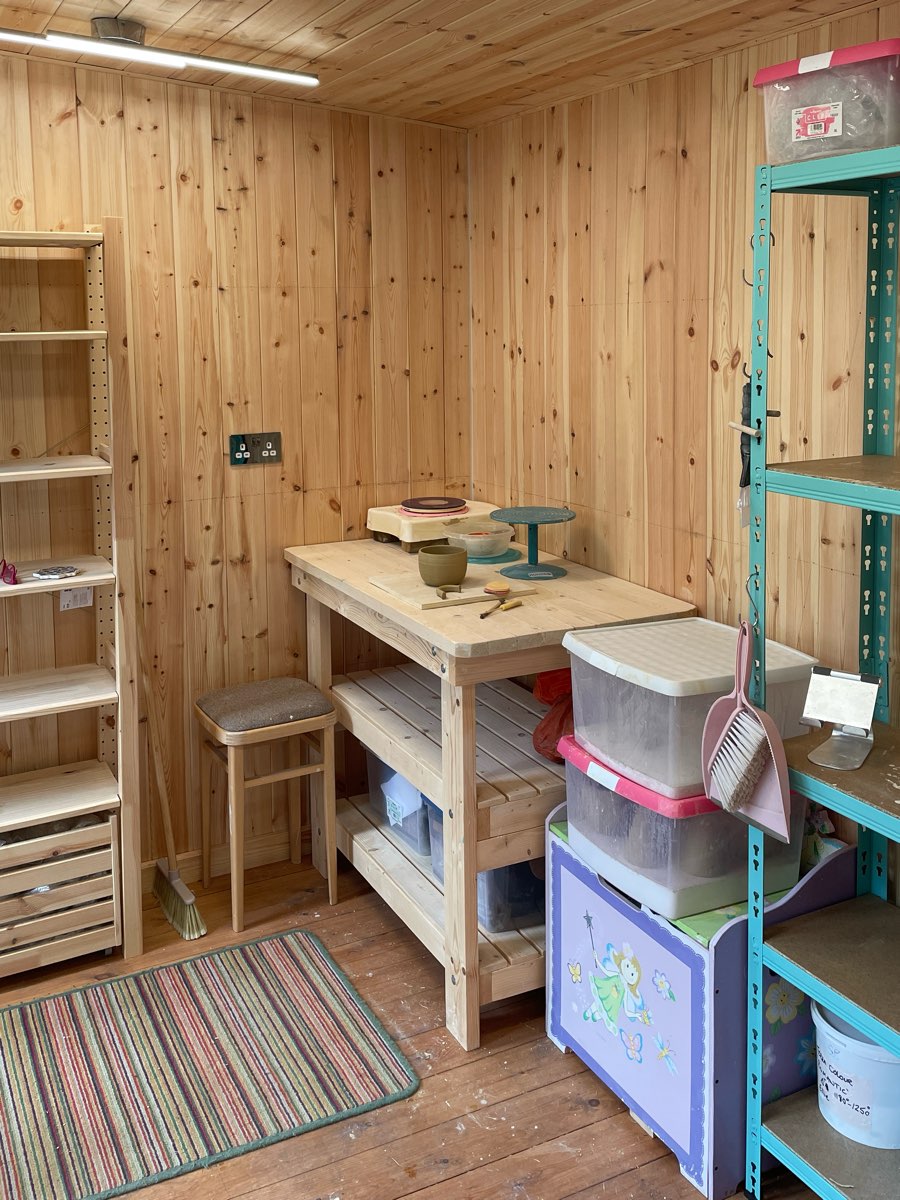 A pine workbench in the corner of the studio with a banding wheel, a freshly thrown pot and trimming tools, beside a stool and shelving